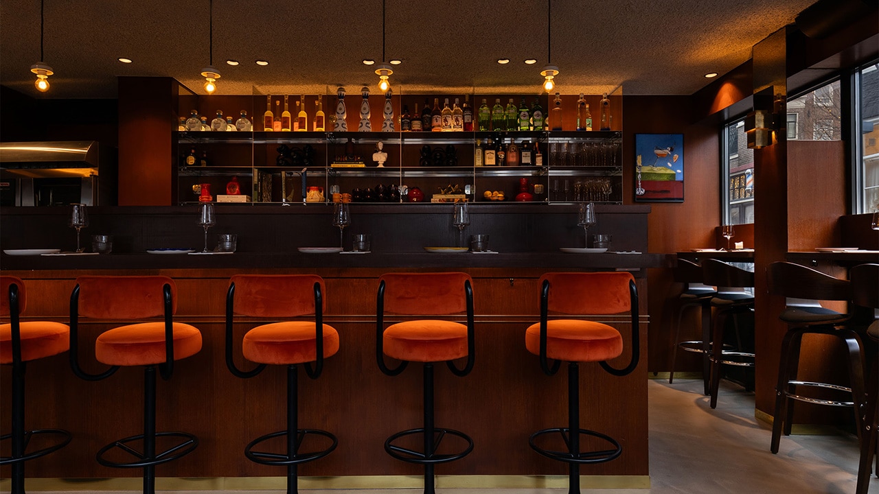 Inside view of Ela Restobar's bar, with bottles decorating the wall, high bar chairs and high window seating ready for dinner service with wine glasses and plates on the tables and bar.